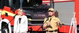 Kobieta w białym stroju oraz strażak w mundurze w kolorze musztardowym stoją przed mikrofonami za nimi stoi sztaluga oraz samochody pożarnicze.