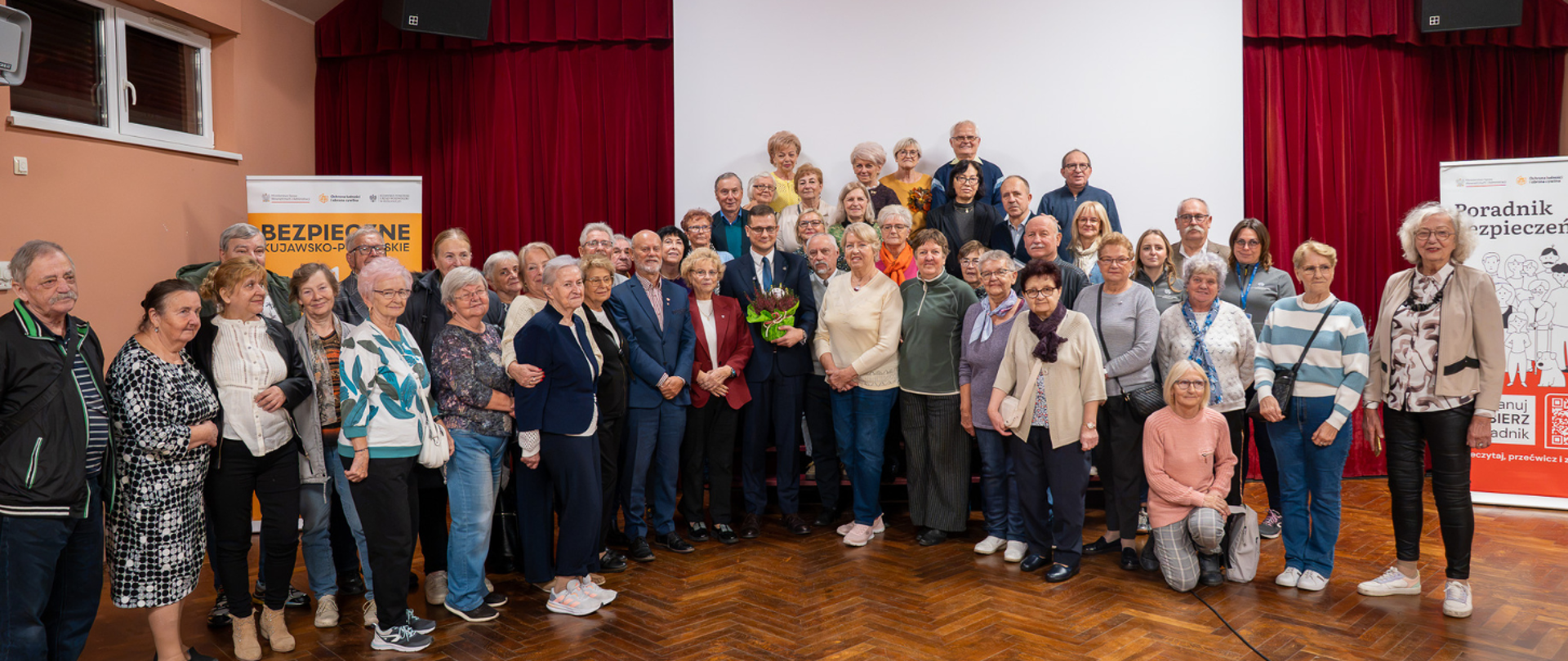 Szerokie pamiątkowe zdjęcie przedstawia liczną grupę seniorów zgromadzonych w sali konferencyjnej. Pośrodku stoi Wojewoda Kujawsko-Pomorski Michał Sztybel w garniturze i okularach, trzymając kwiaty. Po bokach tła ustawiono materiały informacyjne z widocznymi napisami „BEZPIECZNE KUJAWSKO-POMORSKIE” oraz „Poradnik bezpieczeństwa”.