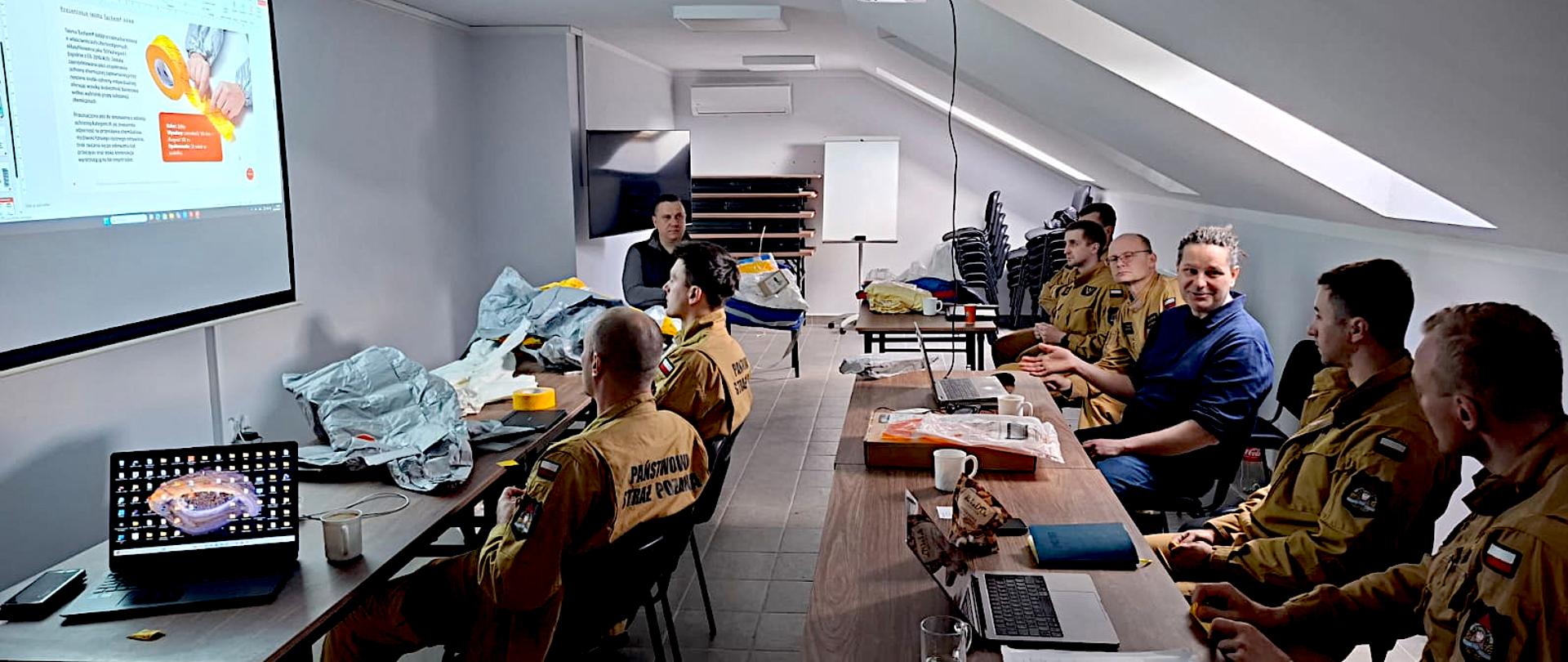 Zdjęcie przedstawia grupę strażaków uczestniczących w szkoleniu z zakresu ratownictwa chemicznego, odbywającym się w sali konferencyjnej na poddaszu. Wzdłuż dwóch długich, równoległych stołów siedzi kilku mężczyzn. Większość z nich ma na sobie piaskowe mundury służbowe z napisem „Państwowa Straż Pożarna” na plecach oraz polskimi flagami na ramionach. Jeden z mężczyzn, ubrany w cywilny ciemny sweter, siedzi bokiem i zdaje się aktywnie uczestniczyć w dyskusji. Na przedniej ścianie znajduje się duży ekran projekcyjny, na którym wyświetlany jest dokument. Obok ekranu, na stole, leżą rozłożone srebrne kombinezony. Na stołach przed strażakami znajdują się otwarte laptopy, notatniki, telefony komórkowe oraz kubki z napojami. W głębi sali widać ustawione w stosy krzesła, białą tablicę (flipchart) oraz klimatyzator.