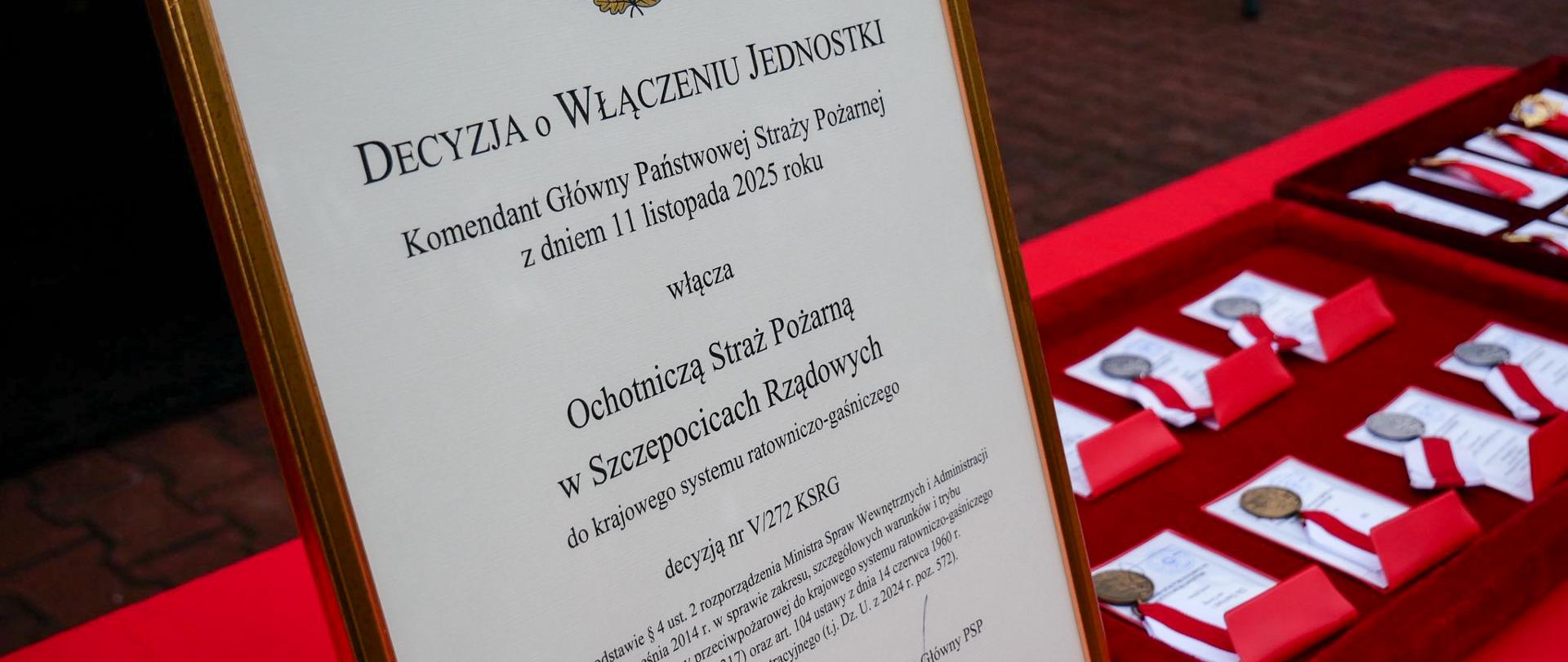 Zbliżenie na oprawiony dokument w języku polskim. Nagłówek to "DECYZJA O WŁĄCZENIU JEDNOSTKI" Państwowej Straży Pożarnej, która włącza Ochotniczą Straż Pożarną w Szczepocicach Rządowych do Krajowego Systemu Ratowniczo-Gaśniczego (KSRG). W prawym dolnym rogu widoczny podpis Komendanta Głównego PSP, nadbryg. Wojciecha Kraczka. Obok leżą medale w czerwonych welurowych pudełkach.