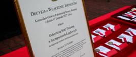 Zbliżenie na oprawiony dokument w języku polskim. Nagłówek to "DECYZJA O WŁĄCZENIU JEDNOSTKI" Państwowej Straży Pożarnej, która włącza Ochotniczą Straż Pożarną w Szczepocicach Rządowych do Krajowego Systemu Ratowniczo-Gaśniczego (KSRG). W prawym dolnym rogu widoczny podpis Komendanta Głównego PSP, nadbryg. Wojciecha Kraczka. Obok leżą medale w czerwonych welurowych pudełkach.