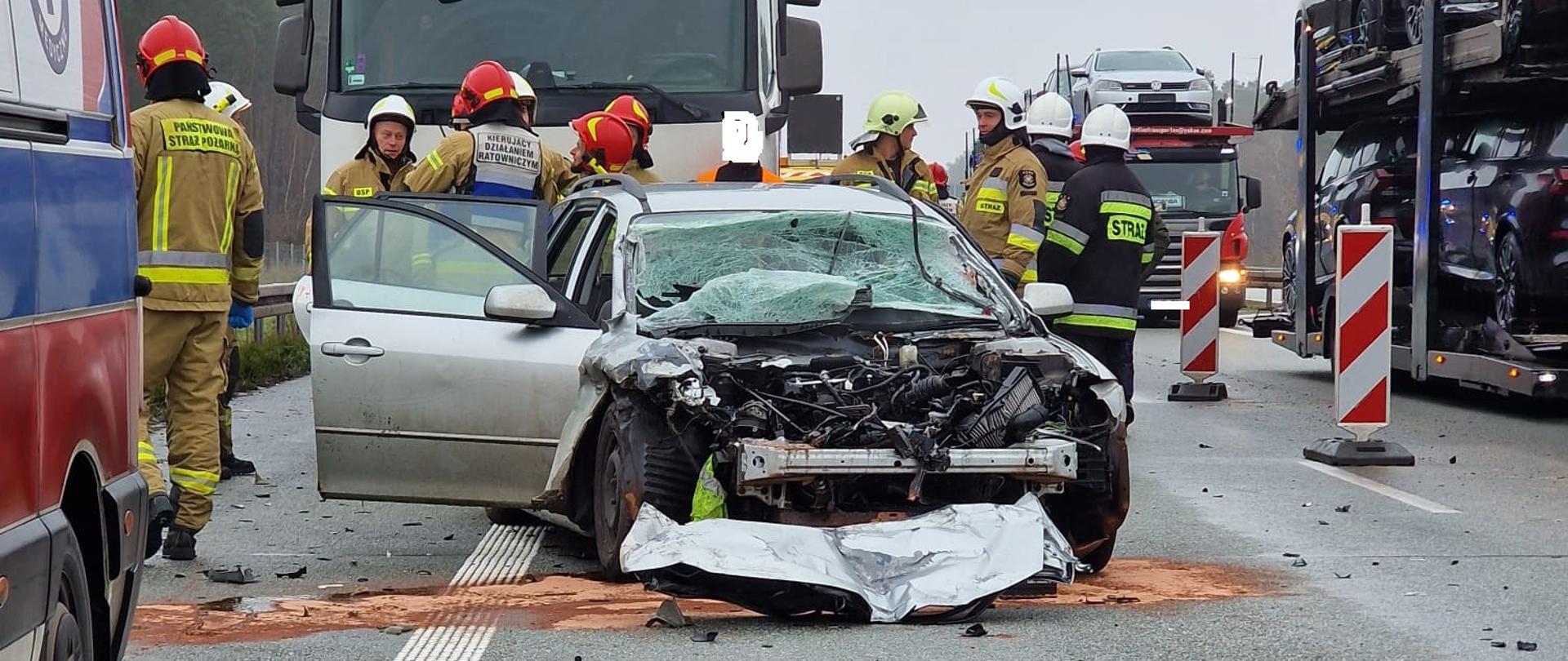 Uszkodzony przód samochodu osobowego wokół strażacy oraz inne samochody