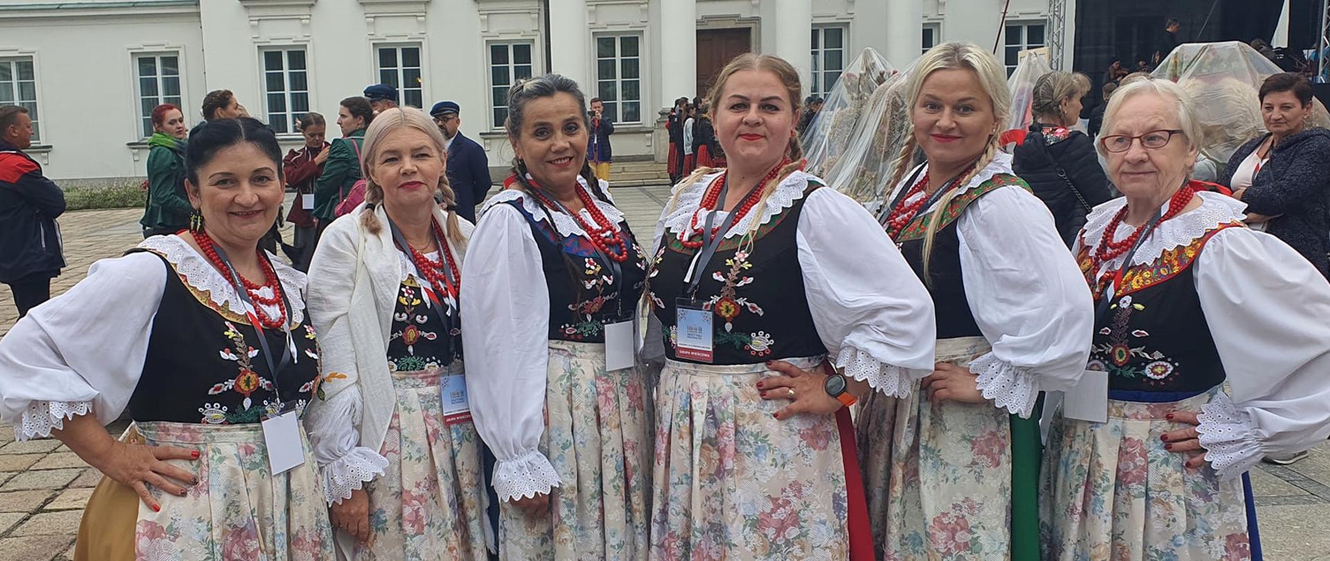 6 pań w strojach ludowych na tle Pałacu Prezydenckiego