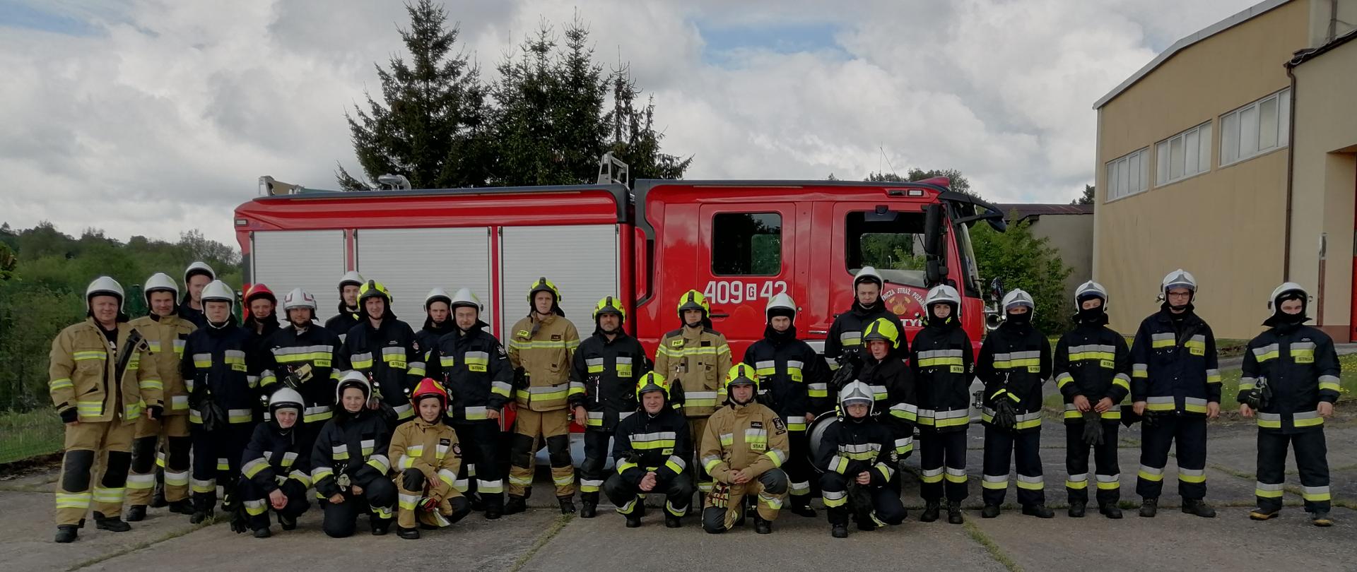 Na zdjęciu znajdują się strażacy ratownicy OSP ubrani w umundurowanie specjalne, na głowach maja hełmy ochronne. Za strażakami ustawiony jest wóz strażacki ratowniczo-gaśniczy.