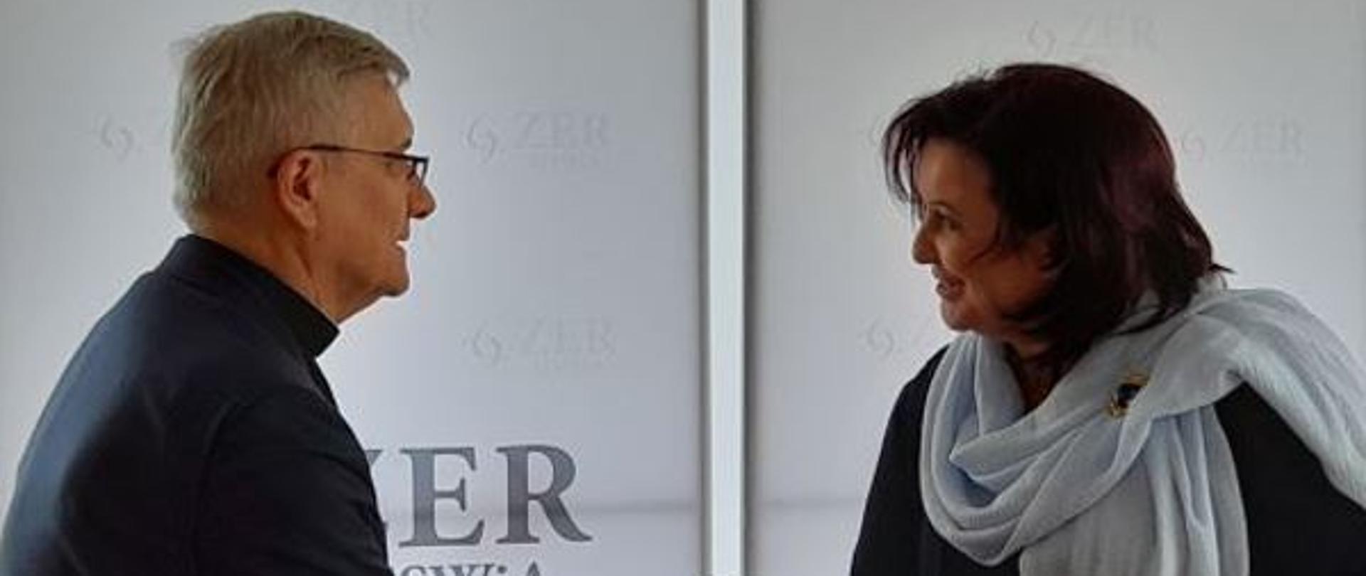 Na zdjęciu jest Pani Małgorzata Zdrodowska, Dyrektor Zakładu Emerytalno-Rentowego Ministerstwa Spraw Wewnętrznych i Administracji oraz ks. prof. dr hab. Ryszard Czekalski, Rektor Uniwersytetu Kardynała Stefana Wyszyńskiego w Warszawie, którzy podają sobie prawe dłonie, a w lewych trzymają czerwone teczki.