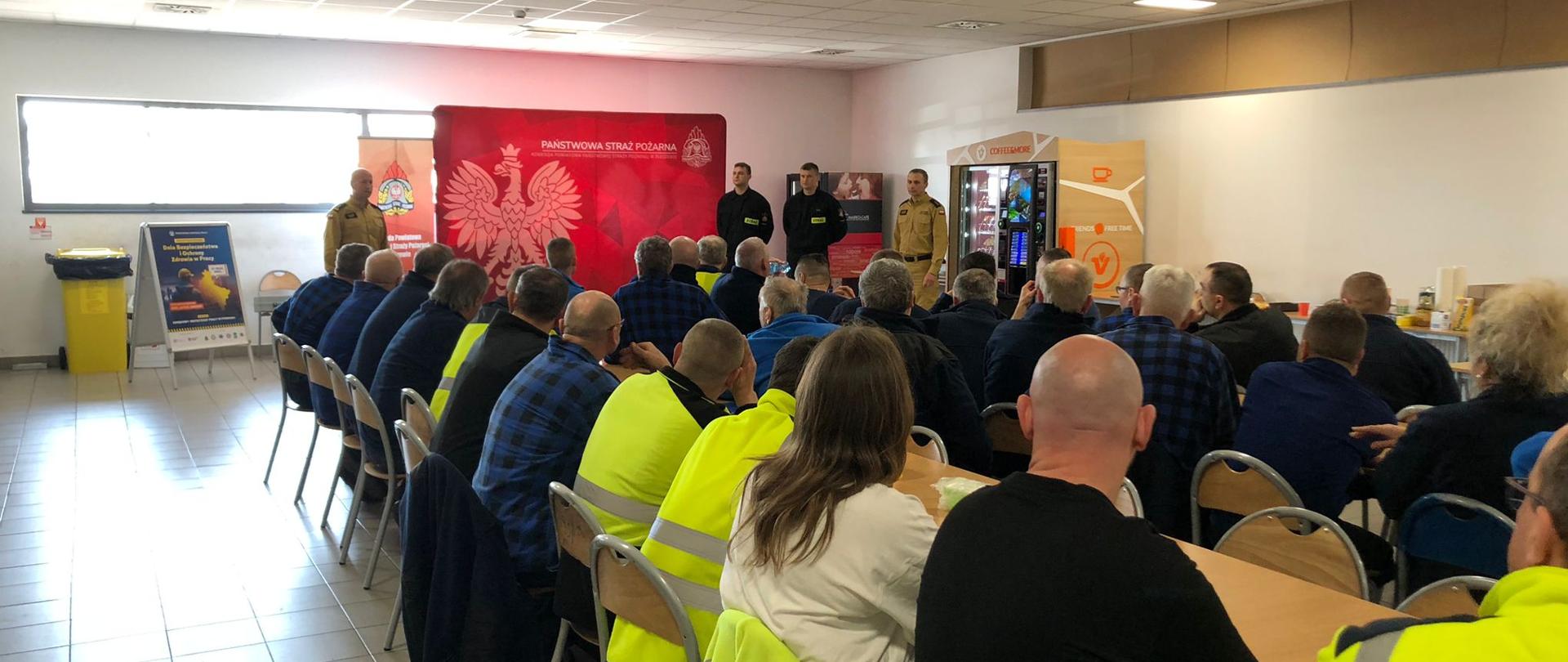 Grupa osób siedzi przy stole w oświetlonej sali, przed nimi stoi czterech mężczyzn ubranych w mundury straży pożarnej, w tle widoczny czerwony baner PSP