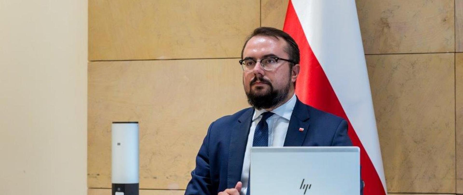 Wiceminister Jabłoński na tle polskiej flagi bierze udział w spotkaniu online