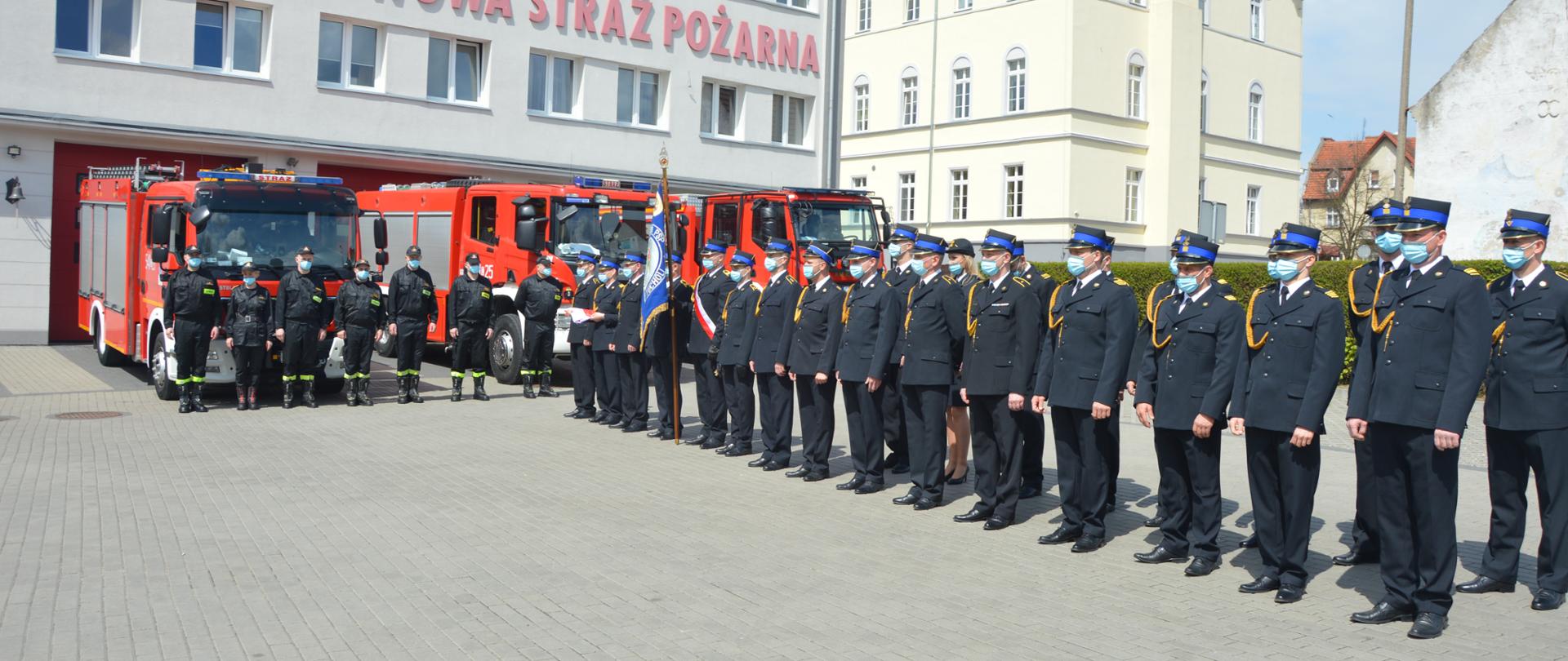 Na zdjęciu widać funkcjonariuszy PSP (w umundurowaniu koszarowym oraz wyjściowym) stojących na placu Komendy Powiatowej PSP w Tucholi. W tle widać budynek komendy, samochody strażackie oraz budynek Szkoły Podstawowej. 