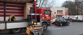 Wózek widłowy ściąga paletę ze strażackiej naczepy podpiętej pod ciągnik siodłowy, w tle parking szpitala, na którym stoją samochody i ambulanse.