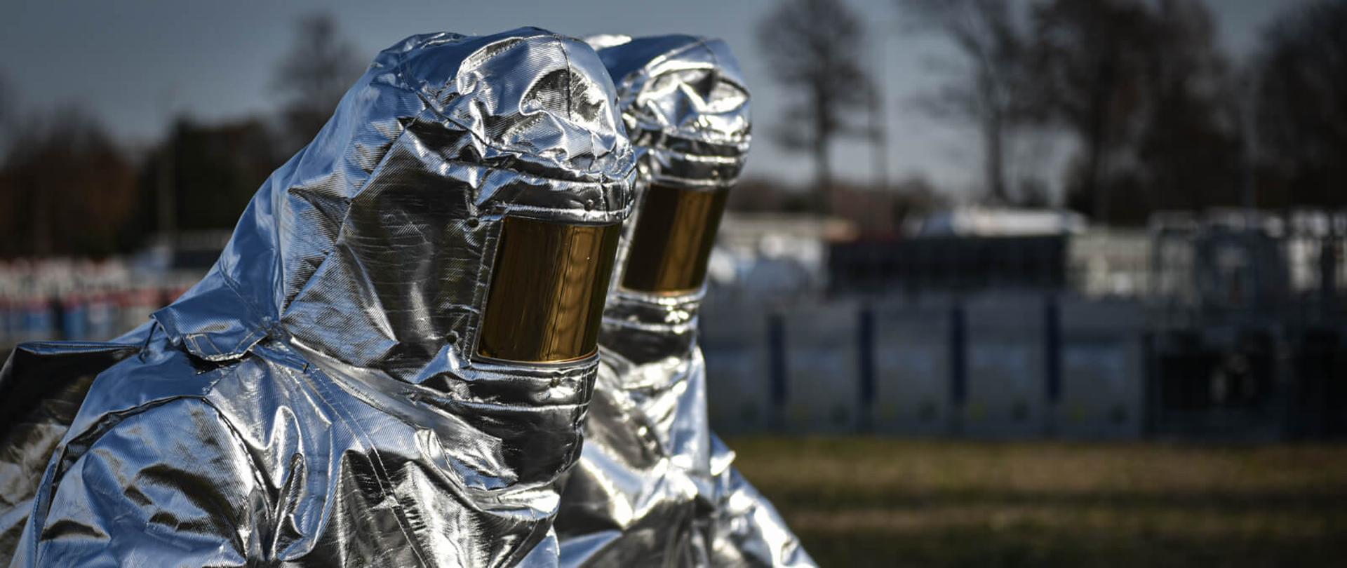 dwóch strażaków w ubraniach żaroodpornych w tle rozmazany obraz terenu zakładu