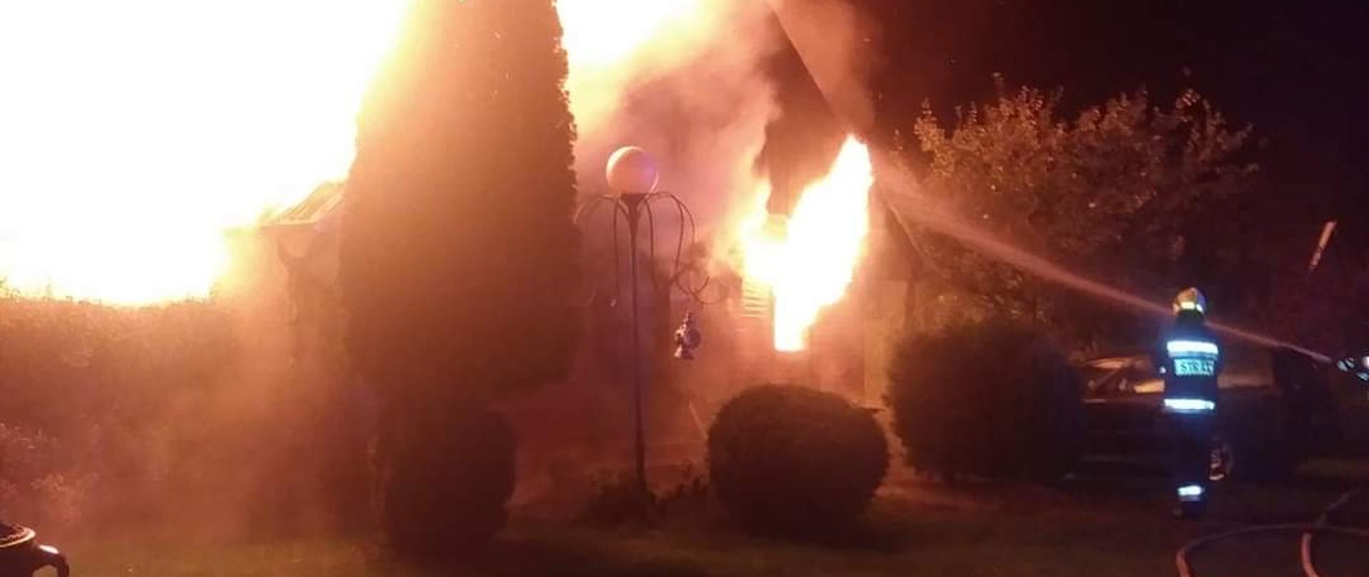 Zdjęcie przedstawia pożar altany w miejscowości Huta Dolna. Pożar w porze nocnej. Altana o konstrukcji drewniej, jednokondygnacyjna z poddaszem. Płomienie obejmują altanę w całości. W dalszym planie po prawej stronie altany w bezpośrednim zagrożeniu usytuowany jest samochód osobowy.
Z prawej strony strażacy podają jeden prąd wody na palącą się altanę i bronią pojazd przed pożarem. 