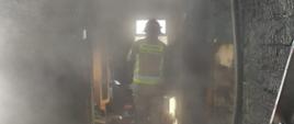 Widoczne na zdjęciu silne zadymienie wewnątrz budynku mieszkalnego, strażak pracujący w środku, widoczne pogorzelisko i zawalony dach