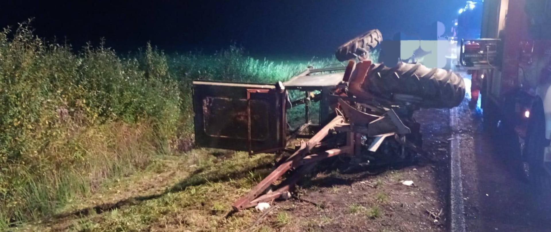 Na zdjęciu widać miejsce wypadku drogowego z udziałem ciągnika rolniczego, który przewrócił się na bok. Maszyna leży na poboczu drogi. Ciągnik jest poważnie uszkodzony — widać zdeformowaną konstrukcję, oderwane elementy ramy i odsłonięte podzespoły. W tle, po prawej stronie kadru, znajdują się pojazdy straży pożarnej z włączonymi światłami ostrzegawczymi. Teren wokół to przydrożny pas zieleni z gęstą roślinnością. Zdjęcie zostało wykonane w nocy, a scena jest oświetlona światłami pojazdów ratunkowych.