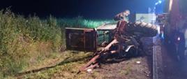 Na zdjęciu widać miejsce wypadku drogowego z udziałem ciągnika rolniczego, który przewrócił się na bok. Maszyna leży na poboczu drogi. Ciągnik jest poważnie uszkodzony — widać zdeformowaną konstrukcję, oderwane elementy ramy i odsłonięte podzespoły. W tle, po prawej stronie kadru, znajdują się pojazdy straży pożarnej z włączonymi światłami ostrzegawczymi. Teren wokół to przydrożny pas zieleni z gęstą roślinnością. Zdjęcie zostało wykonane w nocy, a scena jest oświetlona światłami pojazdów ratunkowych.