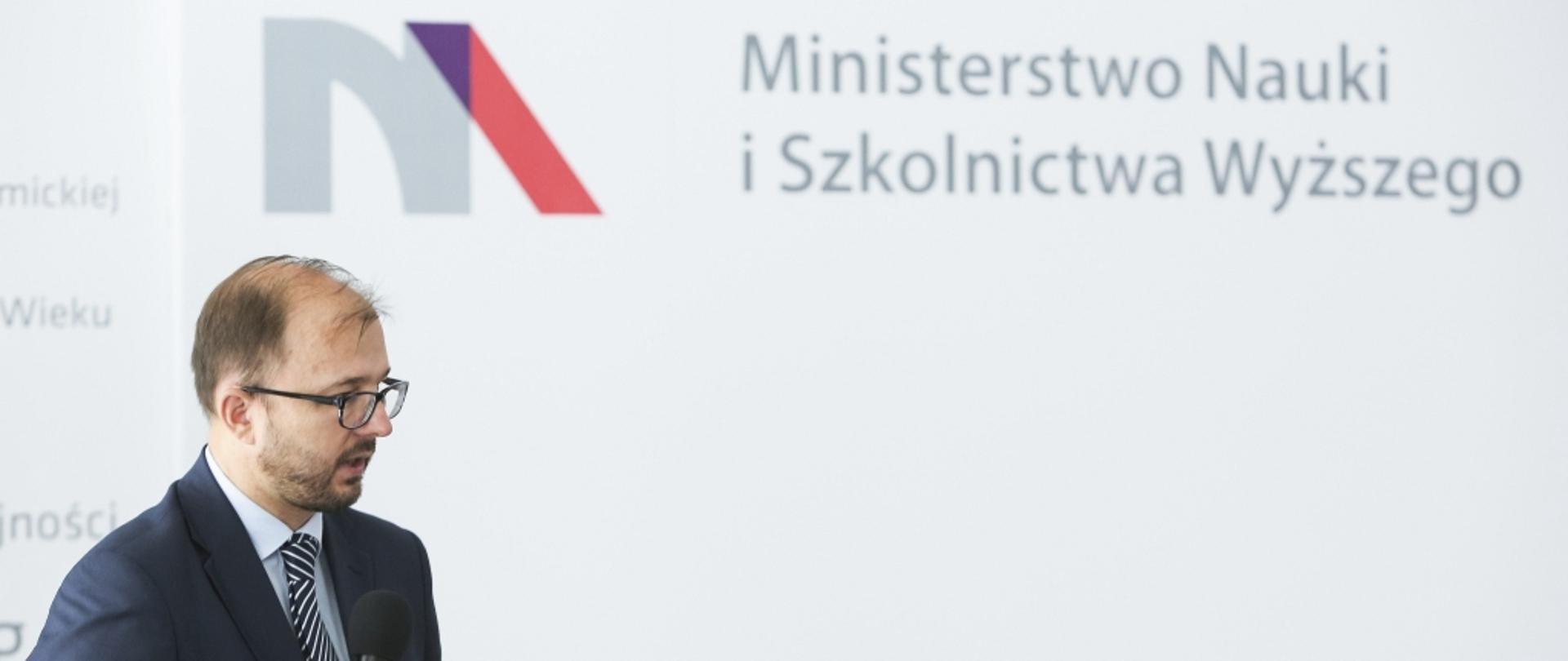 Minister Piotr Dardziński przemawia do mikrofonu, w tle ściana z logiem MNiSW