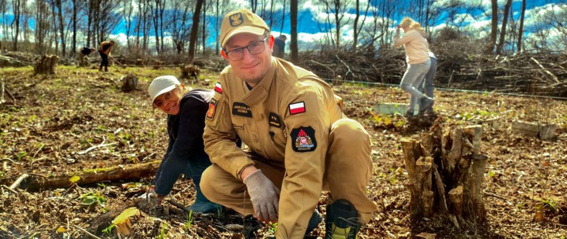 Zdjęcie przedstawia funkcjonariusza PSP podczas sadzenia drzewek wraz z innymi ludźmi. 