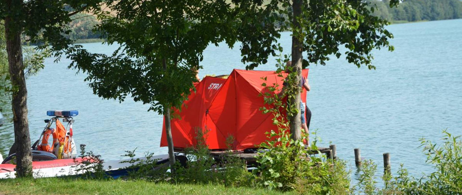 Pomost na którym ustawiono parawan zasłaniający miejsce udzielania pomocy podjętemu z wody poszkodowanemu mężczyźnie. Z lewej strony pomostu znajduje się łódź strażacka.