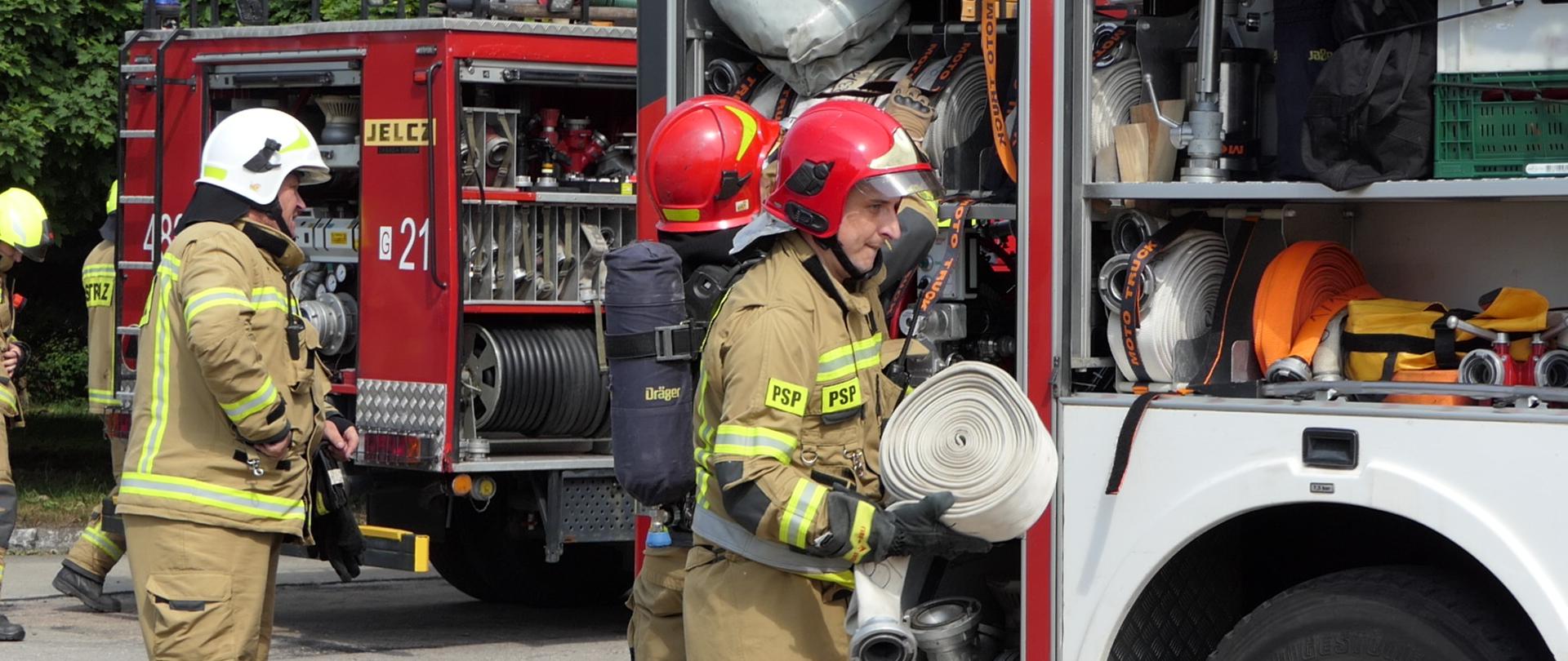 Fotografia przedstawia strażaków rozwijających węże pożarnicze podczas ćwiczeń na tle wozów strażackich.