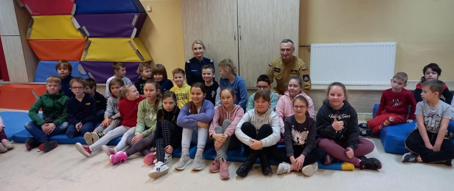Na zdjęciu znajdują się dzieci siedzące na podłodze oraz strażak i policjantka
