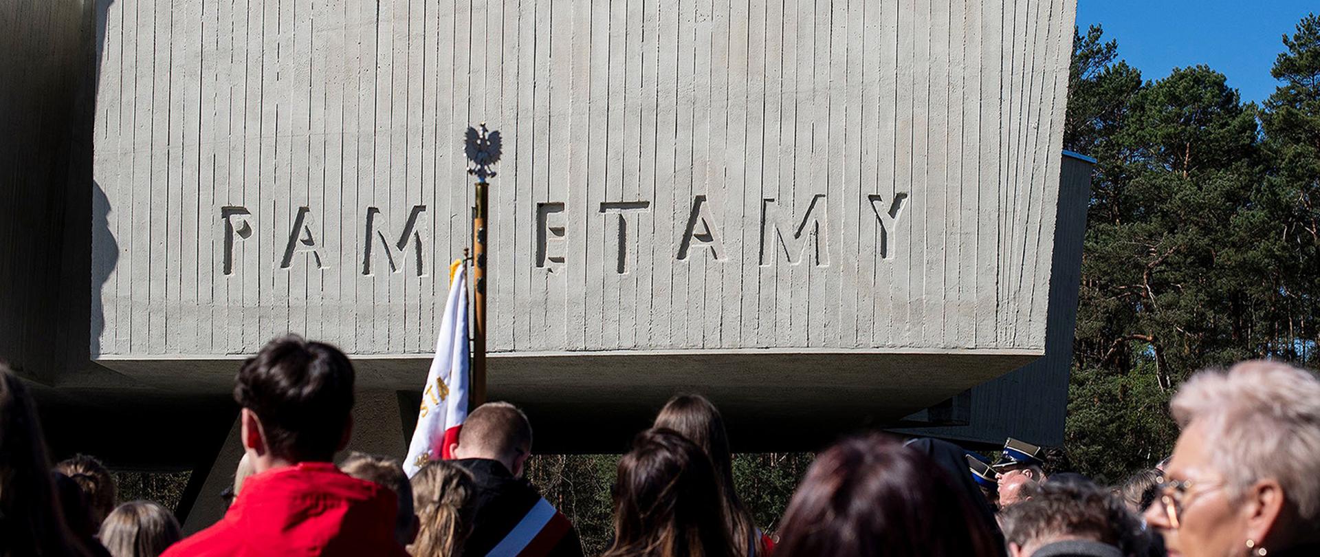 Zdjęcie przedstawia fragment nowoczesnego, betonowego monumentu o pionowej fakturze. Na bocznej ścianie wykuty jest duży, minimalistyczny napis drukowanymi literami: „PAMIĘTAMY” (litera „I” jest zasłonięta przez drzewiec sztandaru). W dolnej części kadru widoczne są czubki głów zgromadzonego tłumu oraz polski orzeł wieńczący jeden ze sztandarów, co nadaje zdjęciu perspektywę żabią i podkreśla ogrom pomnika.