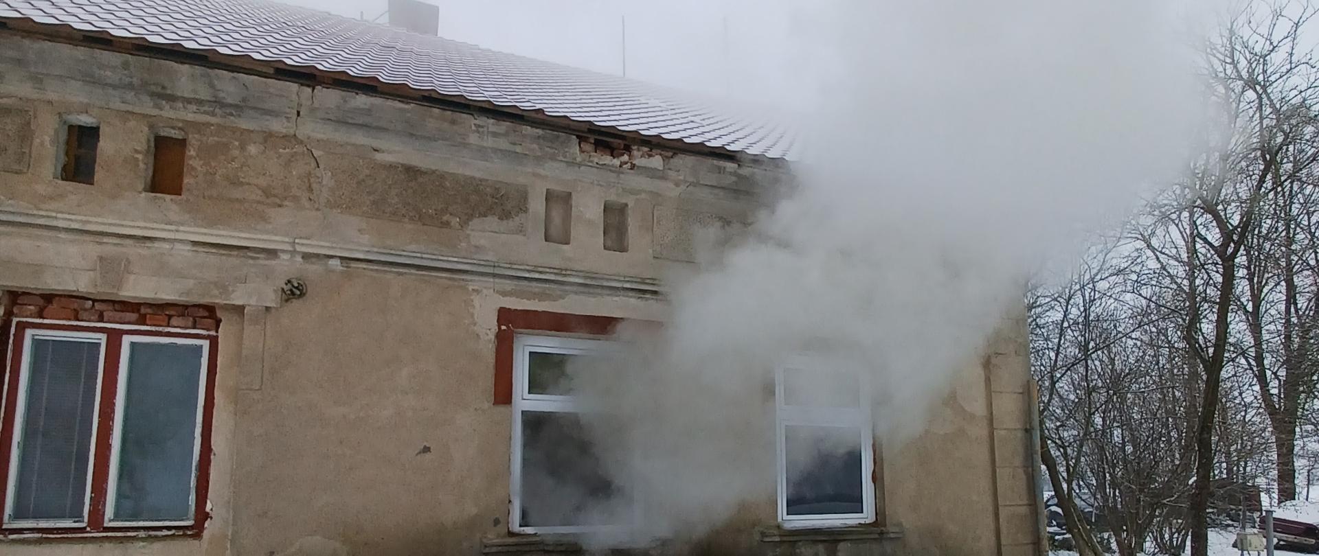 Z budynku mieszkalnego przez otwarte okno wydobywa si dym. Przed budynkiem znajduje się sterta desek.
