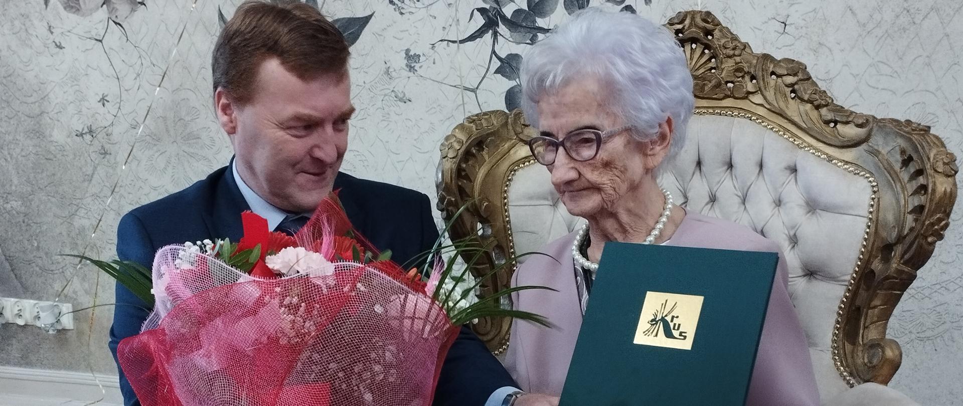 Elegancka starsza kobieta siedzi na zdobionym fotelu. Lekko się uśmiecha , pochylona w kierunku mężczyzny w garniturze , który przyklęknął obok z bukietem kwiatów i ozdobną zieloną teczką z logo KRUS.