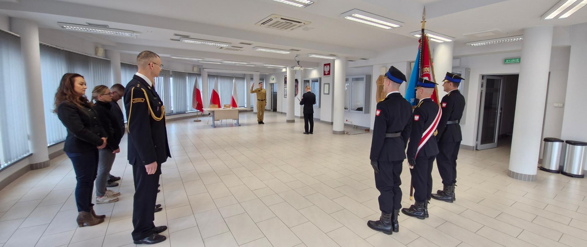 Na sali konferencyjnej znajdują się strażacy oraz osoby cywilne. Strażacy ubrani w mundury wyjściowej z wyjątkiem jednego strażaka który znajduje się w umundurowaniu służbowym w kolorze piaskowym. Na sali znajduje się sztandar w tle flagi Polski przed którymi strażak w ubraniu w kolorze piaskowym składa przysięgę.