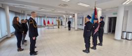 Na sali konferencyjnej znajdują się strażacy oraz osoby cywilne. Strażacy ubrani w mundury wyjściowej z wyjątkiem jednego strażaka który znajduje się w umundurowaniu służbowym w kolorze piaskowym. Na sali znajduje się sztandar w tle flagi Polski przed którymi strażak w ubraniu w kolorze piaskowym składa przysięgę.