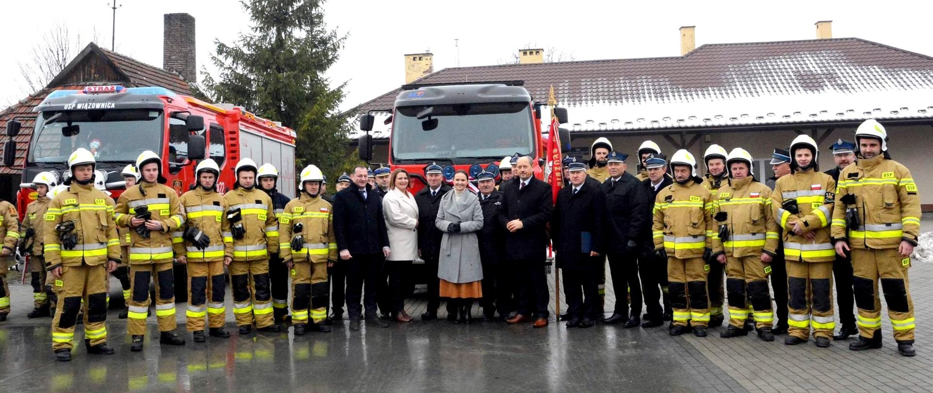 Zdjęcie zrobione na zewnątrz podczas uroczystości przekazania samochodów ratowniczo-gaśniczych. Zdjęcie grupowe uczestników uroczystości. Na zdjęciu strażacy OSP, funkcjonariusze Państwowej Straży Pożarnej oraz osoby cywilne uczestniczące w uroczystości. Za nimi dwa samochody ratowniczo-gaśnicze.
