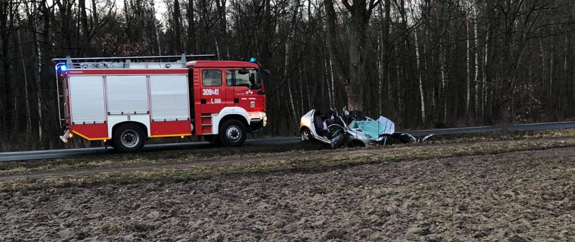 W oddali po lewej stronie na ulicy stoi samochód pożarniczy ciężarowy koloru czerwonego ze srebrnymi skrytkami sprzętowymi należący do OSP Dąbrowa Chełmińska - widać prawy bok auta. W oddali po prawej wrak samochodu marki Peugeot wbity w przydrożne drzewo. Za pojazdami rozciąga się las. 