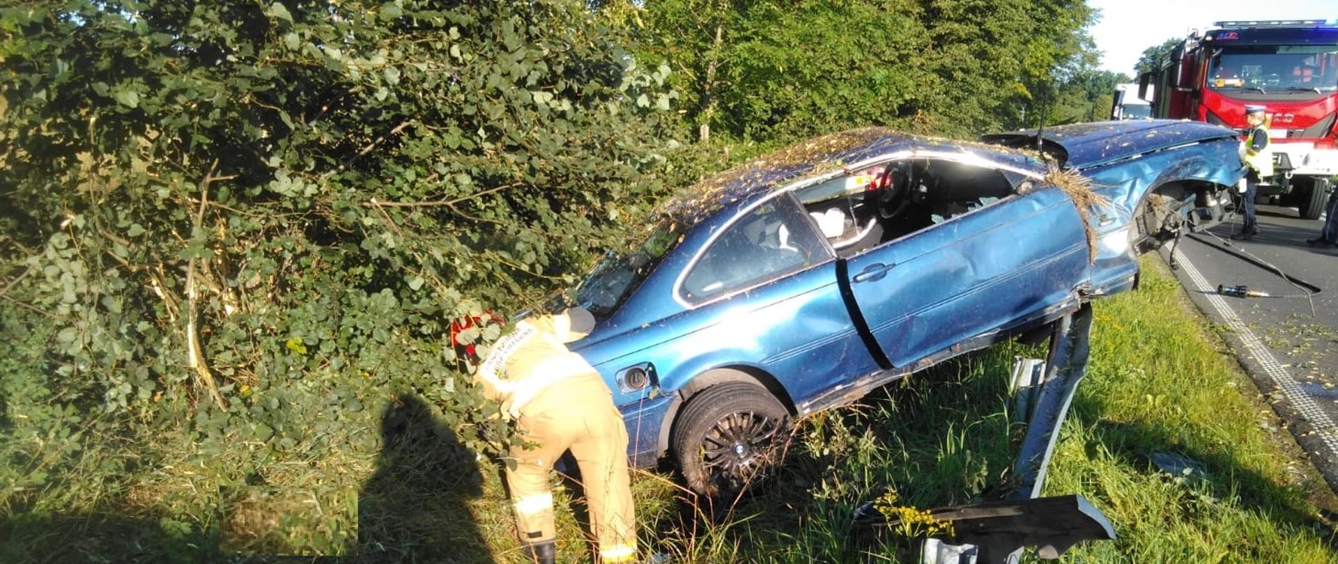 Samochód osobowy znajduje się w rowie oparty o barierę energochłonną. Obok pojazdu stoi strażak, w oddali widać samochód gaśniczy oraz Policjantkę. 