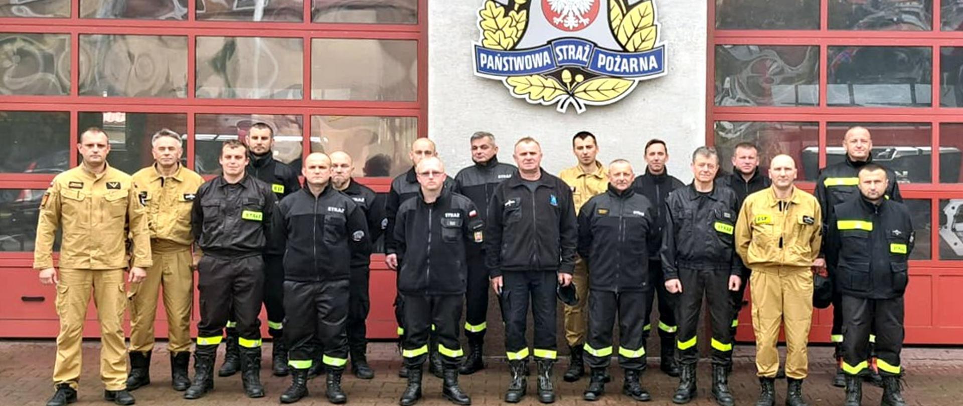 Zdjęcie przedstawia biorących udział w szkoleniu strażaków OSP oraz prowadzącego kurs strażaka PSP. Wszyscy stoją przed garażami JRG.