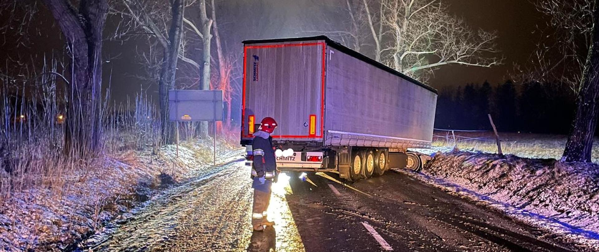 Na zdjęciu widać strażaka stojącego na ulicy za samochodem ciężarowym, który wpadł w poślizg. Strażak jest w ciemnym mundurze i czerwonym hełmie. Na zdjęciu widać naczepę samochodu ciężarowego. W oddali zdjęcia widać drzewa nad naczepą samochodu ciężarowego. Po boku jezdni widać śnieg. Po lewej stronie zdjęcia widać odwrócony znak. 