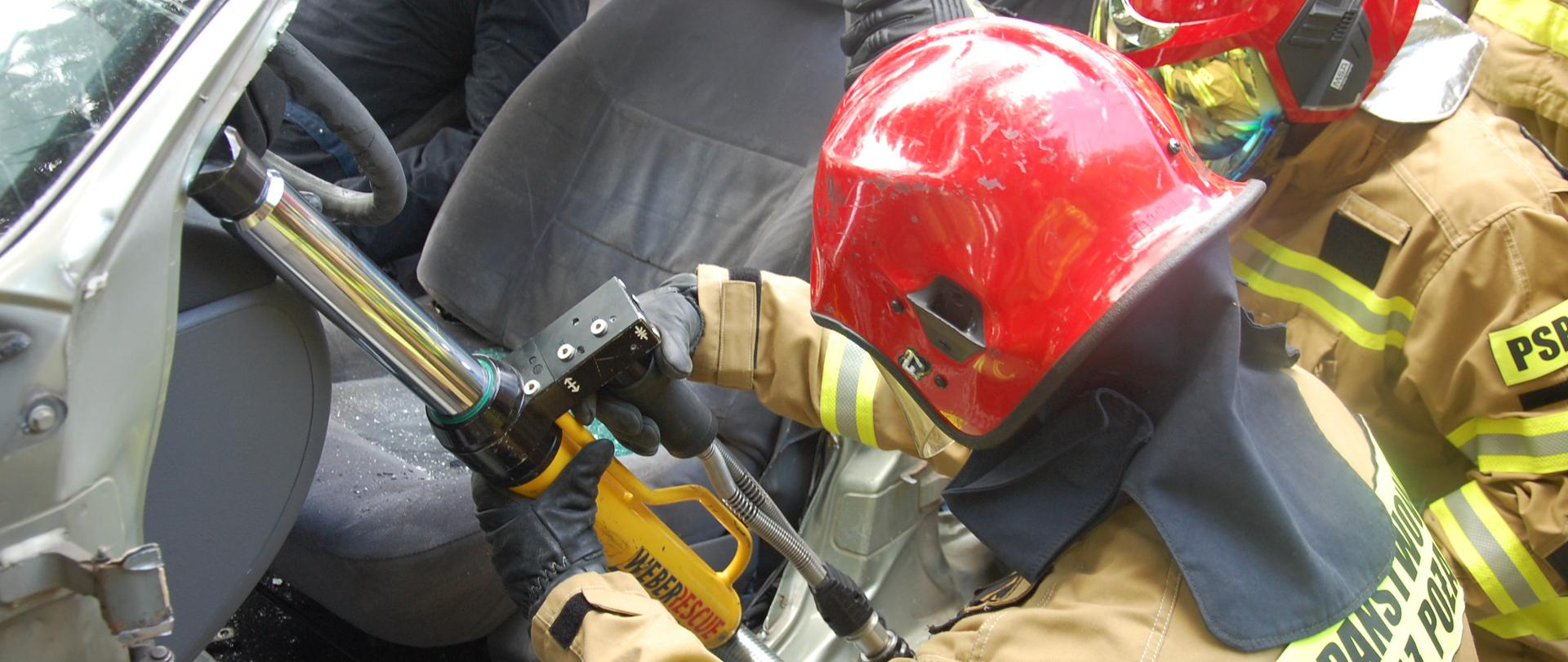 Na zdjęciu dwóch strażaków w ubraniach specjalnych, hełmach oraz rękawicach technicznych podnoszących deskę rozdzielczą za pomocą hydraulicznego rozpieracza kolumnowego umiejscowionego w progu przednich drzwi oraz słupku A pojazdu.