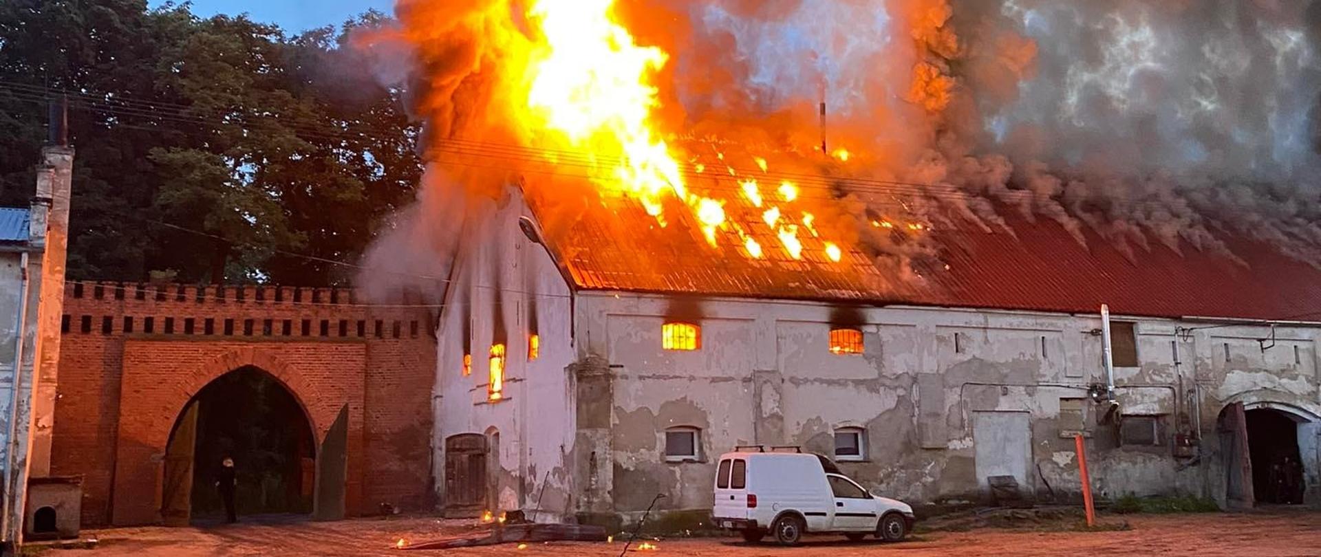 Zdjęcie przedstawia budynek inwentarski objęty pożarem. Ogień wychodzi ponad dach budynku. Przed budynkiem stoi samochód dostawczy.
