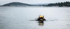 Na zdjęciu strażak nurek ubrany w hełm i skafander podczas wychodzenia z wody na jeziorze Solińskim w tle zapora