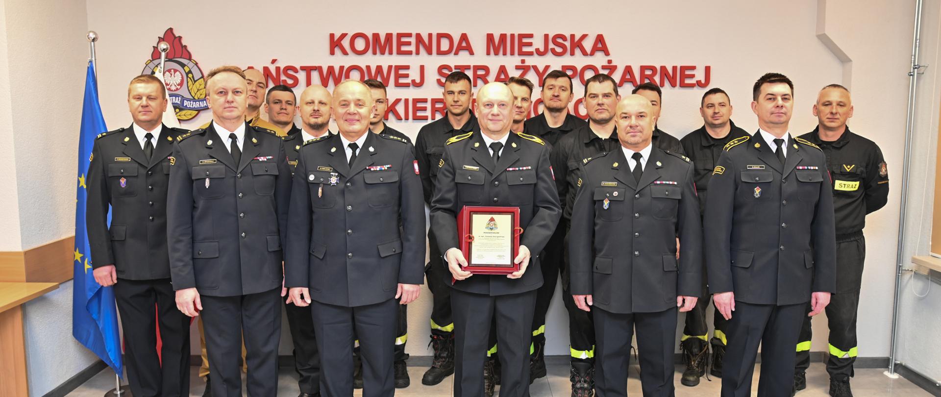 Strażacy w mundurach koloru czarnego stoją na tle napisu koloru czerwonego.