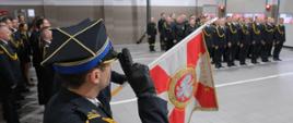 Widok na poczet sztandarowy i strażaków podczas zbiórki w garażu JRG 5. Strażak na pierwszym planie salutuje.