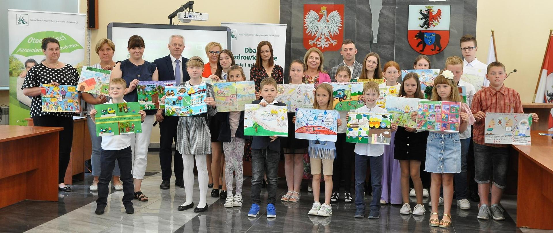 Grupa dzieci z pracami plastycznymi w rękach pozuje do zdjęcia, z tyłu grupa dorosłych osób.