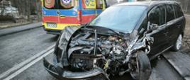 Uszkodzony czarny samochód osobowy po zderzeniu drogowym, z poważnie zniszczonym przodem pojazdu, stoi na jezdni; w tle zaparkowana karetka pogotowia ratunkowego podczas akcji służb.