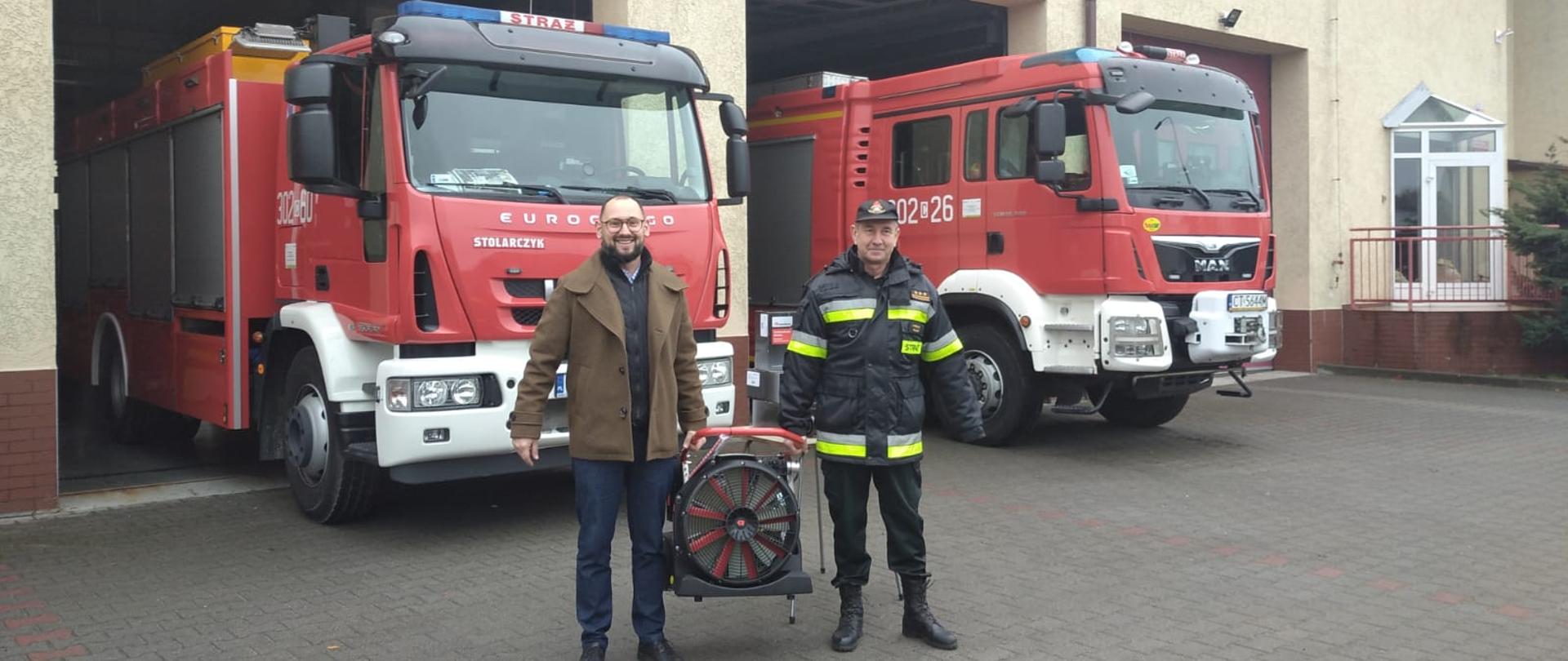 Na pierwszym planie prezes firmy Stena i st. bryg. Janisław Buller Komendant Miejski PSP w Bydgoszczy, wspólnie trzymający wentylator oddymiający, w tle frontowa elewacja JRG2 i przody samochodów pożarniczych wystających z garażu przodem. 