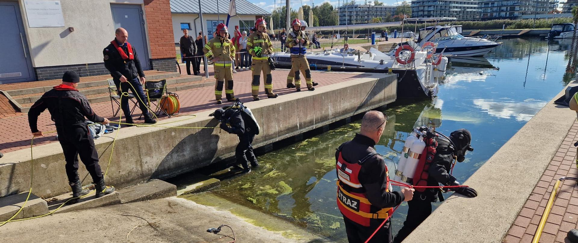Na zdjęciu widać akcję służb ratunkowych przy nabrzeżu portowym. Kilku strażaków i nurków w specjalistycznym sprzęcie bierze udział w działaniach przy wodzie. Jeden z nurków wchodzi do wody, asekurowany przez kolegów za pomocą liny. Inni strażacy obserwują przebieg akcji, część stoi na brzegu, a w tle widać budynek portowy, jachty zacumowane przy kei oraz nowoczesne budynki mieszkalne. Pogoda jest słoneczna, niebo częściowo zachmurzone.