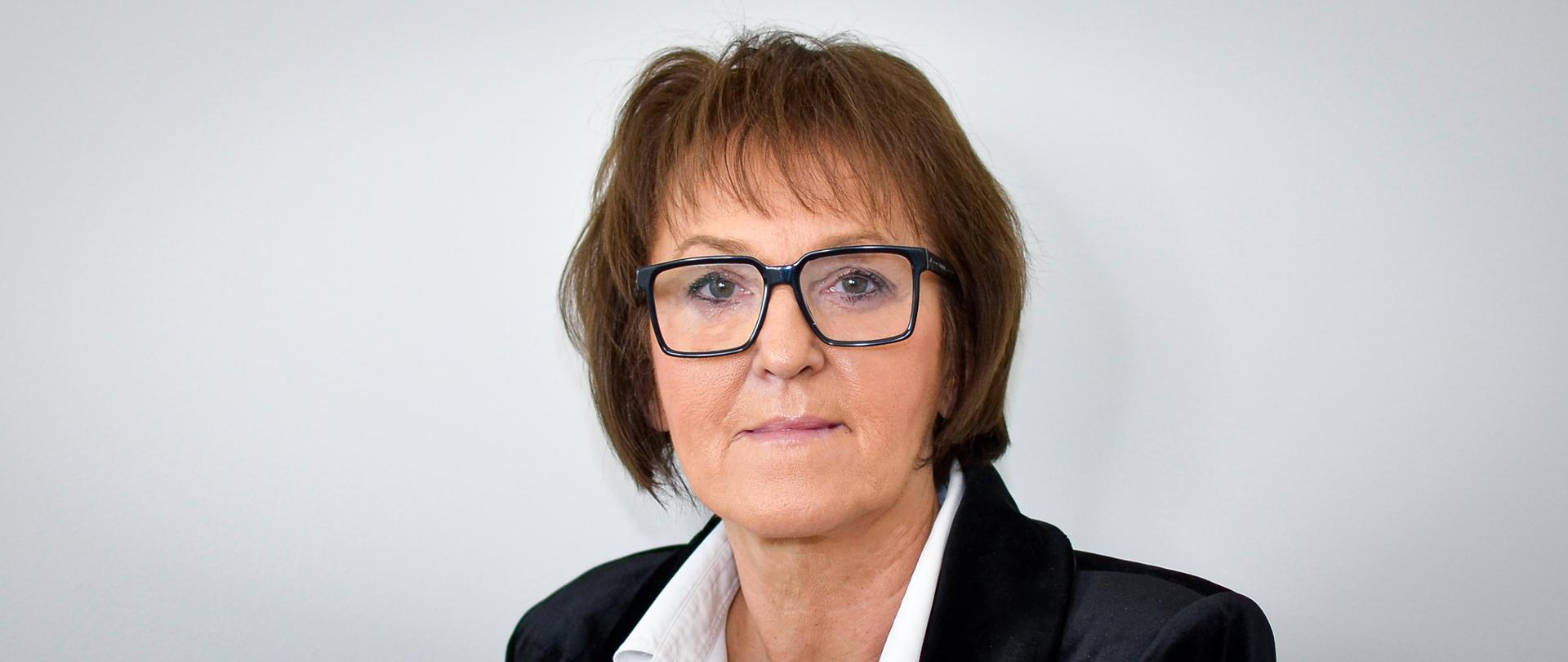 Kobieta w czarnej marynarce, białej bluzce, w okularach, z ciemnymi, krótkimi włosami i grzywką.