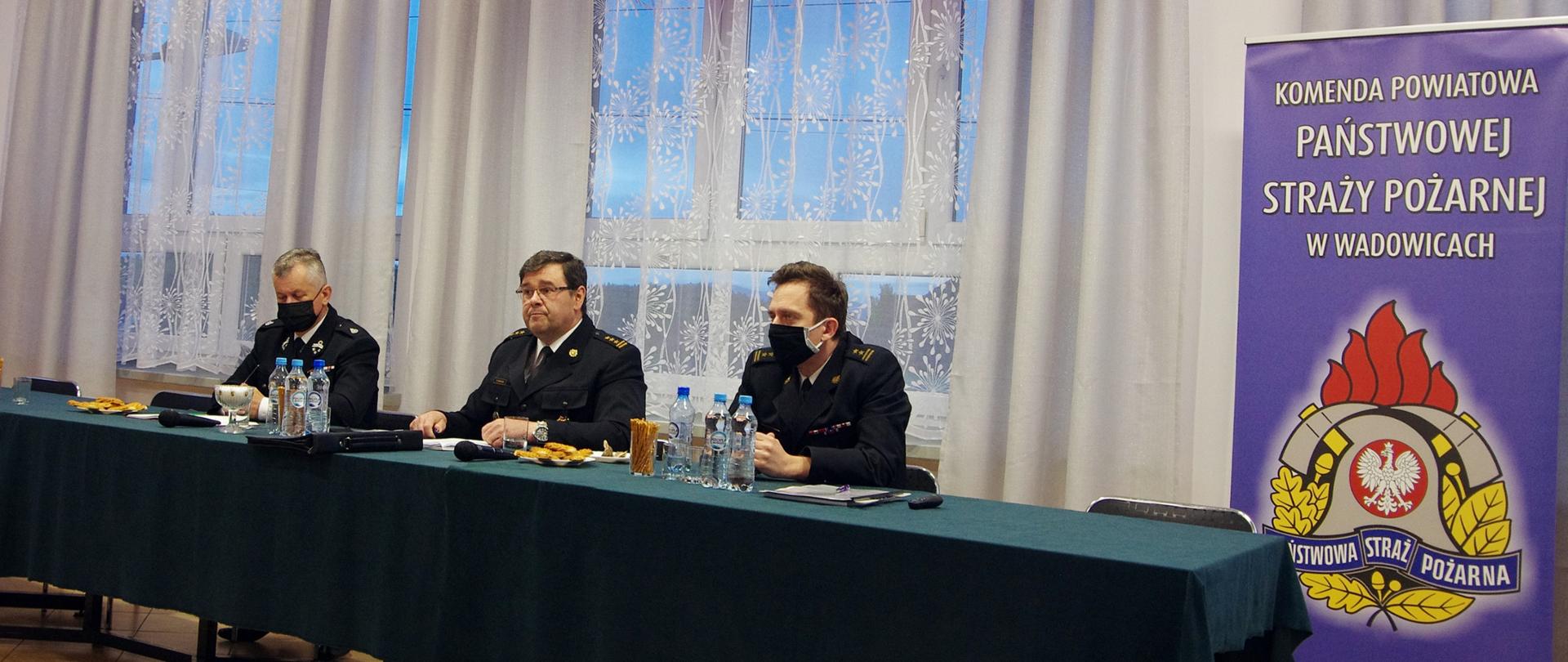 Widok z przodu. Trzech strażaków w czarnych mundurach wyjściowych i maseczkach ochronnych za stołem prezydialnym. Nad nimi widoczna flaga Związku OSP RP. Po prawej stronie baner KP PSP w Wadowicach. 