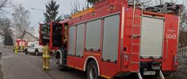 Zdjęcie w ciągu dnia. Na zdjęciu widać pojazd ratowniczo-gaśniczy w kolorze czerwonym, posiada sygnalizację ostrzegawczą. Zdjęcie w pobliżu budynku, w którym doszło do pożaru. Widać ratowników w ubraniu specjalnym.