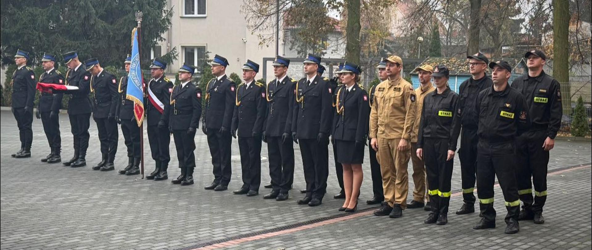 Funkcjonariusze PSP oraz druhowie w ubraniach koszarowych stoją w równym szyku na placu przed jednostką. Po lewej stronie widoczny jest poczet sztandarowy z niebieskim sztandarem. Część strażaków ubrana jest w mundury galowe, pozostali w stroje koszarowe. W tle znajdują się budynki oraz drzewa w jesiennej scenerii.