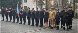 Funkcjonariusze PSP oraz druhowie w ubraniach koszarowych stoją w równym szyku na placu przed jednostką. Po lewej stronie widoczny jest poczet sztandarowy z niebieskim sztandarem. Część strażaków ubrana jest w mundury galowe, pozostali w stroje koszarowe. W tle znajdują się budynki oraz drzewa w jesiennej scenerii.