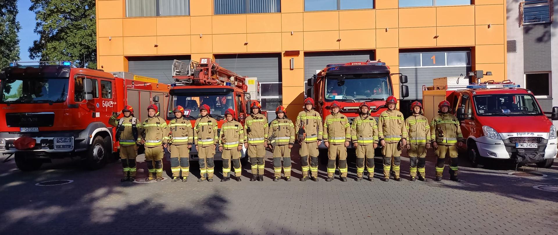 Strażacy ustawieni w rzędzie oddają hołd zmarłemu strażakowi. W tle auta pożarnicze z włączonymi sygnałami świetlno-dźwiękowymi. Z tylu budynki Komendy. Zdjęcie zrobione na placu, na zewnątrz.