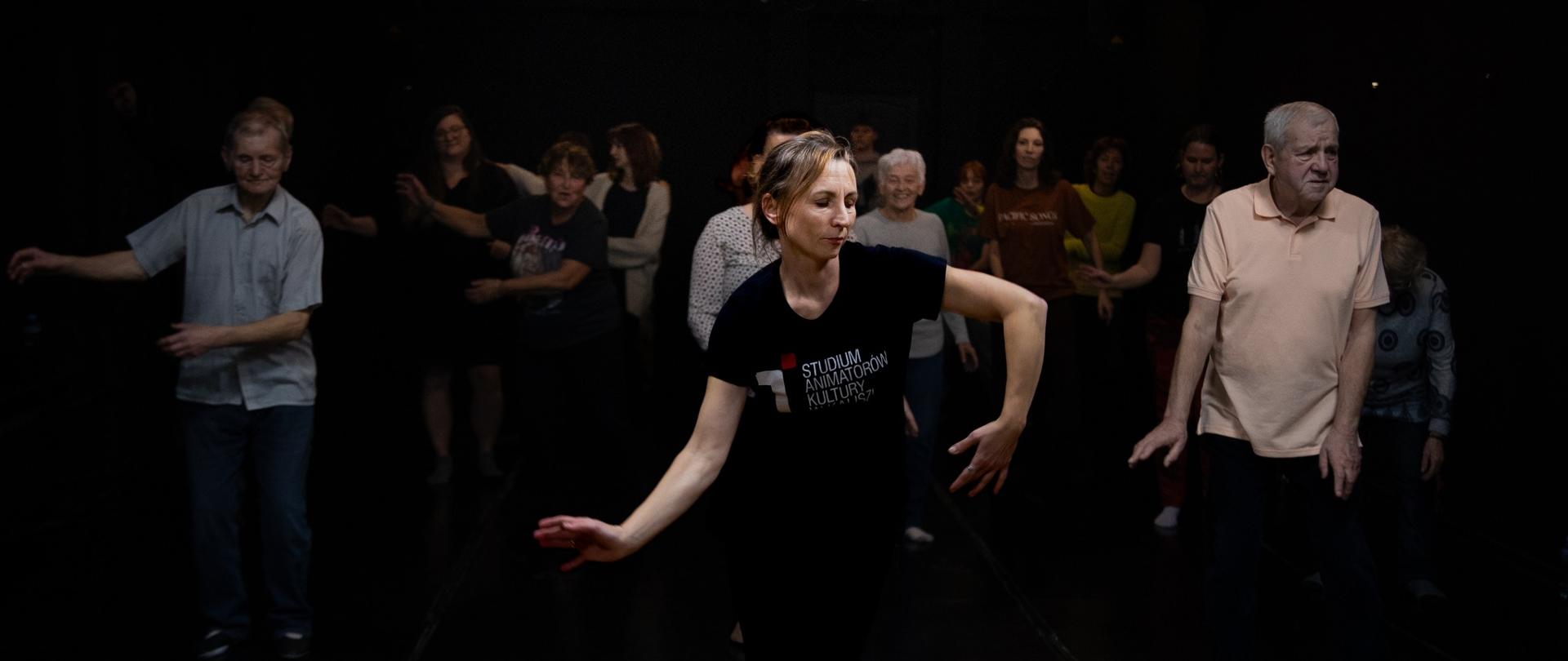 Grupa dorosłych uczestników ćwiczy układ taneczny na ciemnej scenie. Na pierwszym planie widać instruktorkę w czarnej koszulce z napisem „Studium Animatorów Kultury”, która wykonuje dynamiczny ruch rękami. Za nią stoi kilkanaście osób w różnym wieku, odtwarzających te same gesty. Scena jest słabo oświetlona, a podłoga z przodu ma jaśniejsze, drewniane wykończenie.