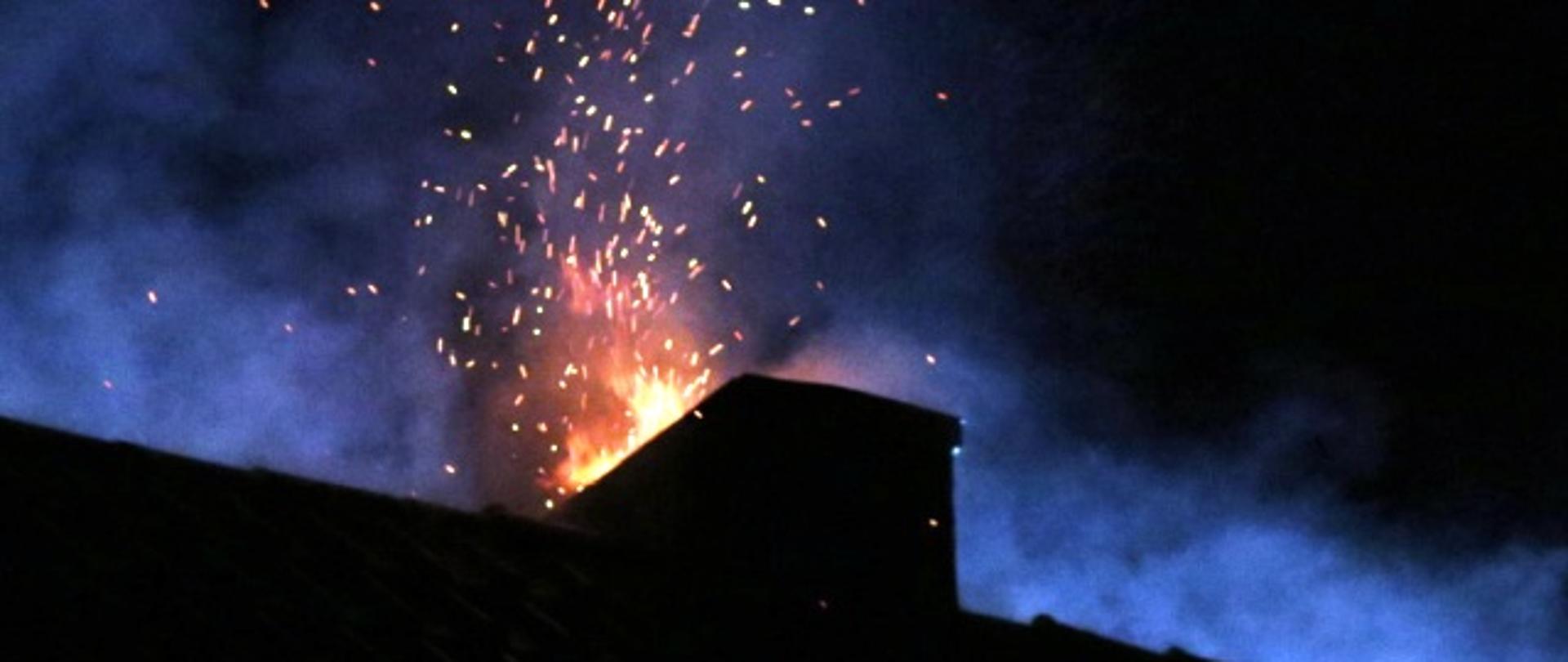 Zdjęcie przedstawia palące się sadze w kominie.
W tle dach budynku i niebo.
